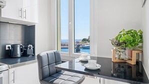 A modern kitchen with a black countertop, a white chair, and a view of the sea through large windows. - Sunlight Properties - Capitaine 54 - 1 Bed rental apartment, balcony, port view (Nice)