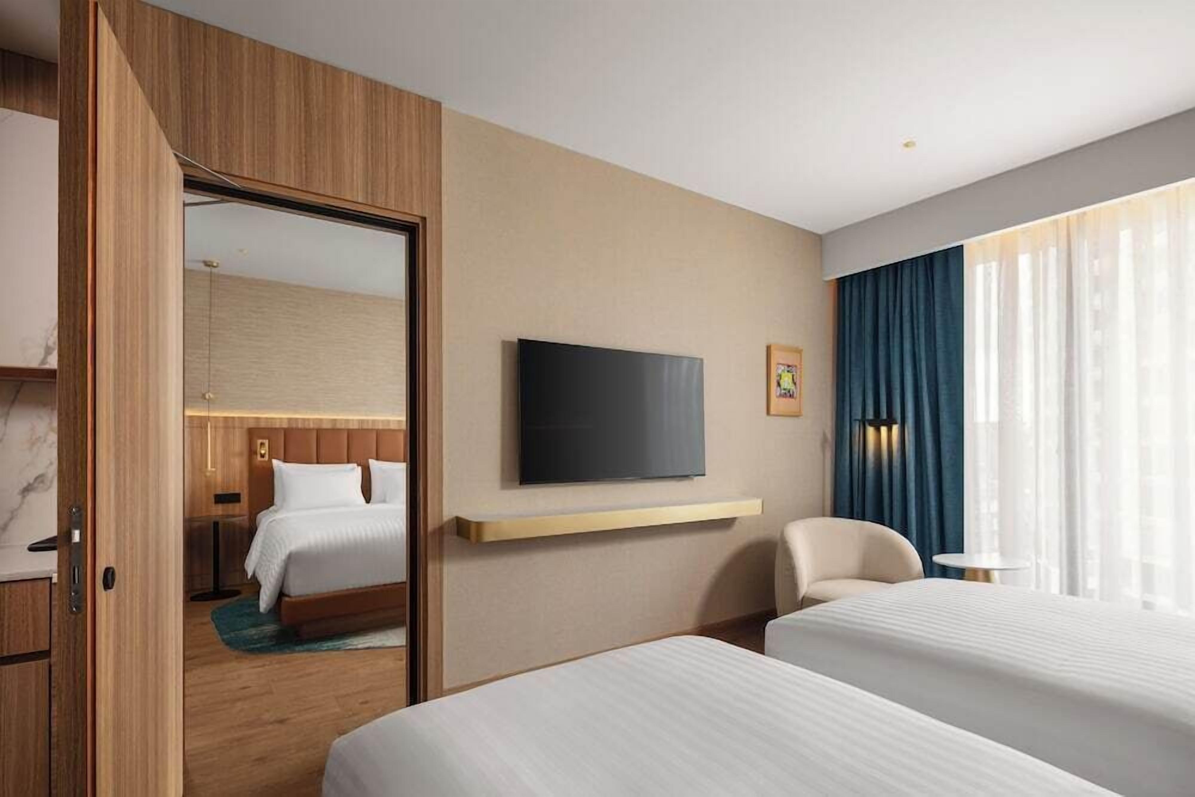 A hotel room with a bed, a TV mounted on the wall, a chair, and a window with curtains.