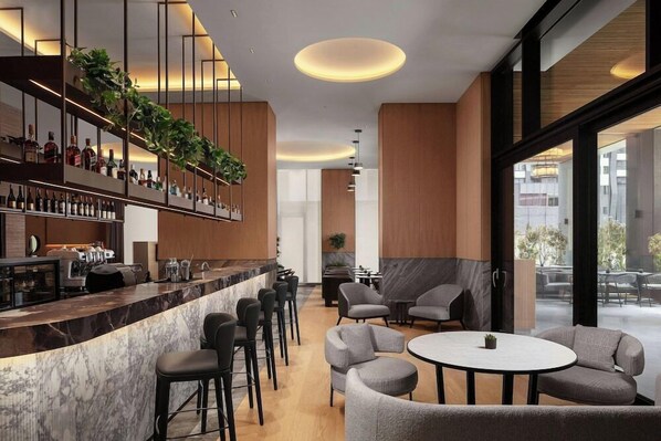 A modern bar area with a marble countertop, bar stools, and a variety of bottles. There are seating areas with round tables and chairs, and a large window offering a view of the outside. - Courtyard by Marriott Prishtina (Pristina)
