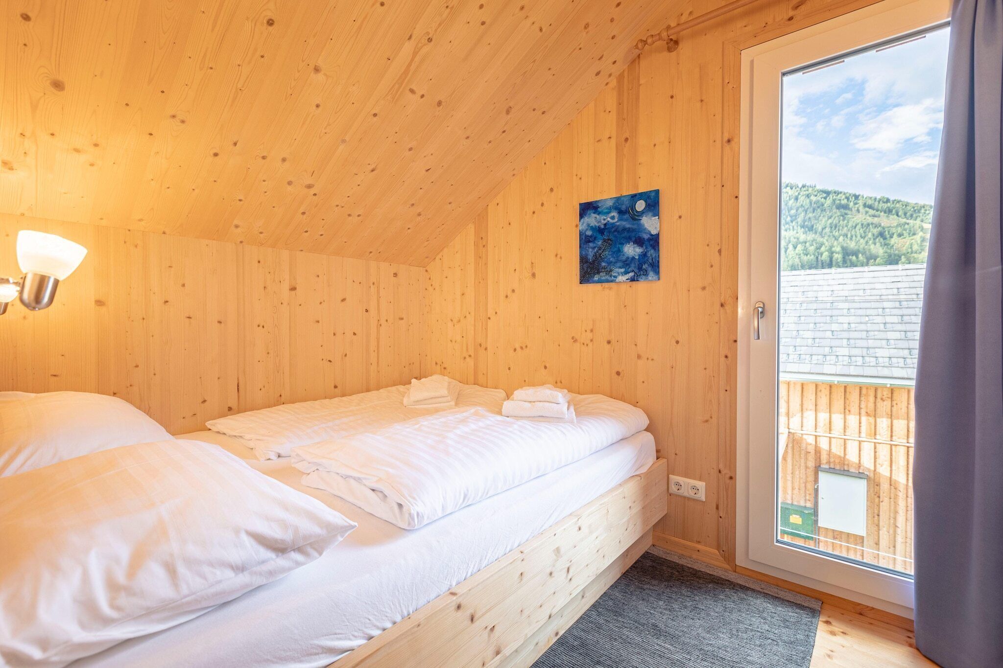A wooden cabin room with a bed, a painting on the wall, and a view of a forested hillside through a door.