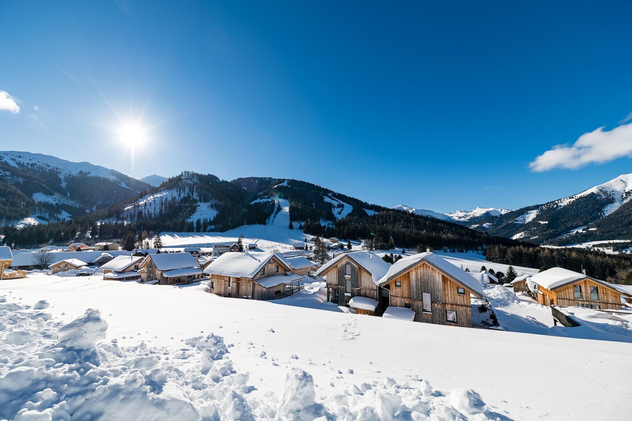 Ein verschneites Dorf mit Holzhütten, einem klaren Himmel und Bergen im Hintergrund.