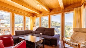 A wooden interior with a sofa, red chairs, and a table. Large windows offer views of a mountainous landscape.
