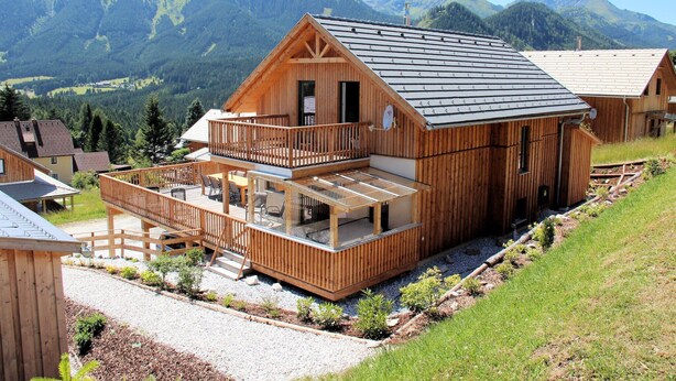 A wooden chalet with a balcony, surrounded by a gravel path and small plants, set against a mountainous backdrop.