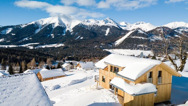 A wooden cabin with a snow-covered roof, surrounded by a snowy landscape and mountains in the background.
