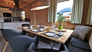 A dining area with a wooden table set for four, surrounded by modern chairs, a large pendant light, and a view of a mountain through a window with curtains.