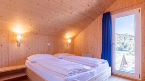A wooden cabin room with two beds, a window with a view of the outdoors, and wall-mounted lights.