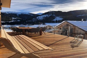 Une terrasse en bois avec une table et des chaises, d’où l’on peut admirer un paysage enneigé avec des montagnes et un chalet.