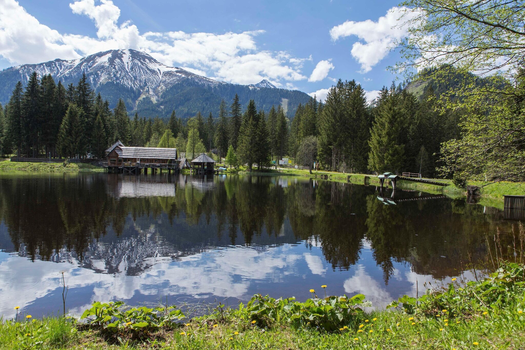 Un cadre paisible au bord d’un lac, avec un chalet en bois et un belvédère au bord de l’eau, le tout entouré d’une végétation luxuriante et dominé, au loin, par une montagne aux sommets enneigés.