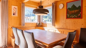 A wooden dining area with a large table and chairs, a hanging light fixture, and a painting on the wall.