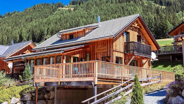 A wooden chalet with a balcony, a stone foundation, and a mountain backdrop.