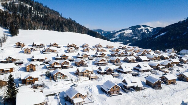 Ein schneebedecktes Dorf mit Holzhäusern, umgeben von Bergen und Kiefern.