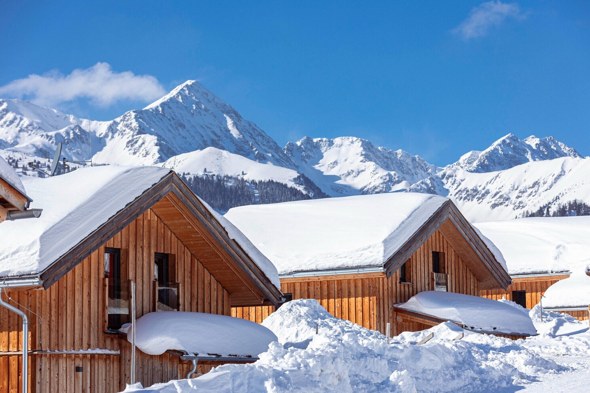 Holzhütten mit schneebedeckten Dächern vor einer Kulisse schneebedeckter Berge unter blauem Himmel.