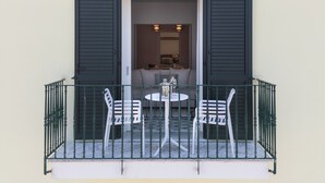 Ein Balkon mit Tisch und zwei Stühlen, eine Tür mit Rollläden und ein Blick in einen Innenraum mit Sofa und Tisch.