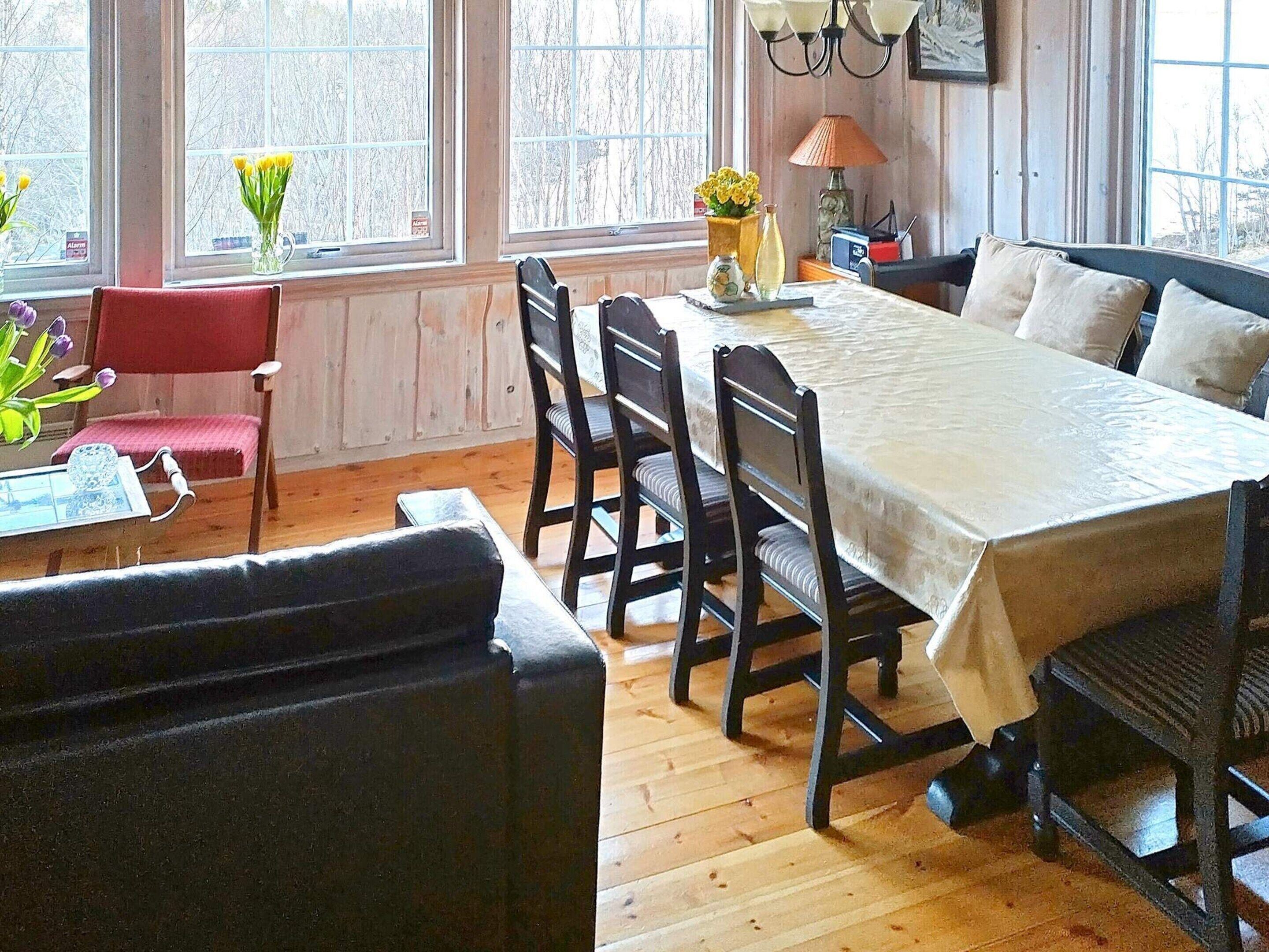 A dining area with a wooden table, four chairs, a red chair, a small table with a vase of flowers, and a lamp on a side table.