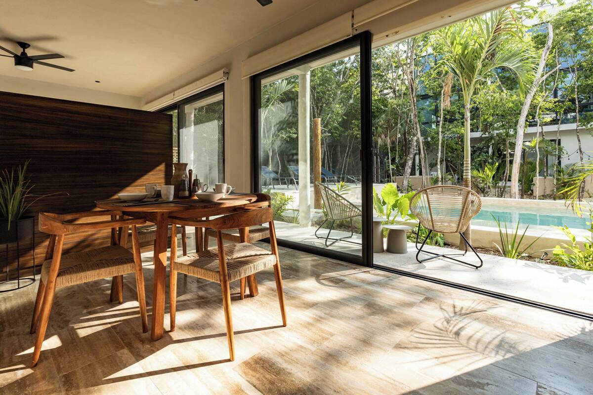 Een moderne eethoek met een houten tafel en stoelen, grote ramen en uitzicht op een zwembad en groen.