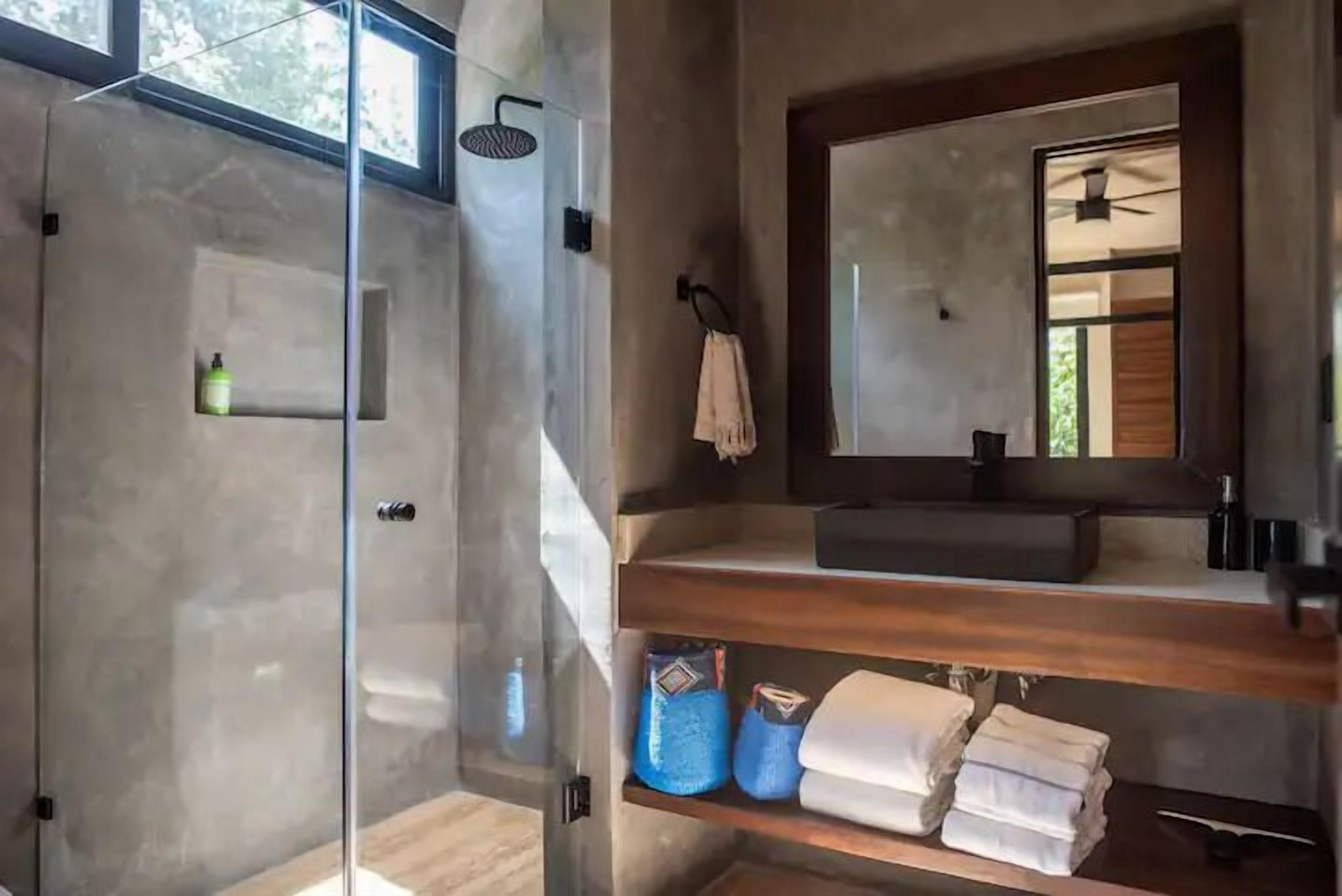 A modern bathroom with a glass-enclosed shower, a wooden vanity with a mirror, and shelves with towels and toiletries.