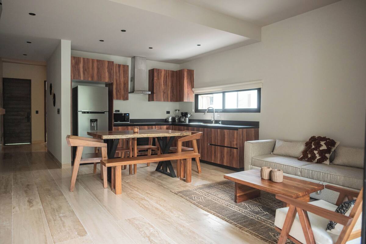 Een moderne keuken met houten kasten, een eethoek met een houten tafel en stoelen, een bank en een groot raam voor natuurlijk licht.
