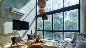 A modern living room with a large window, a flat-screen TV, a sofa, a chair, and a coffee table. - BT Homes - Wellness Jungle (Tulum)