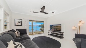 A modern living room with a grey sofa, a black rug, a ceiling fan, a TV, and a view of a lake through a sliding door. - Yaringaa by HolidayCo. (HARDYS BAY)
