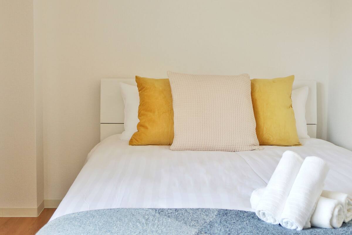 A neatly made bed with white bedding, two pillows (one yellow, one beige), and two rolled white towels on a blue blanket.