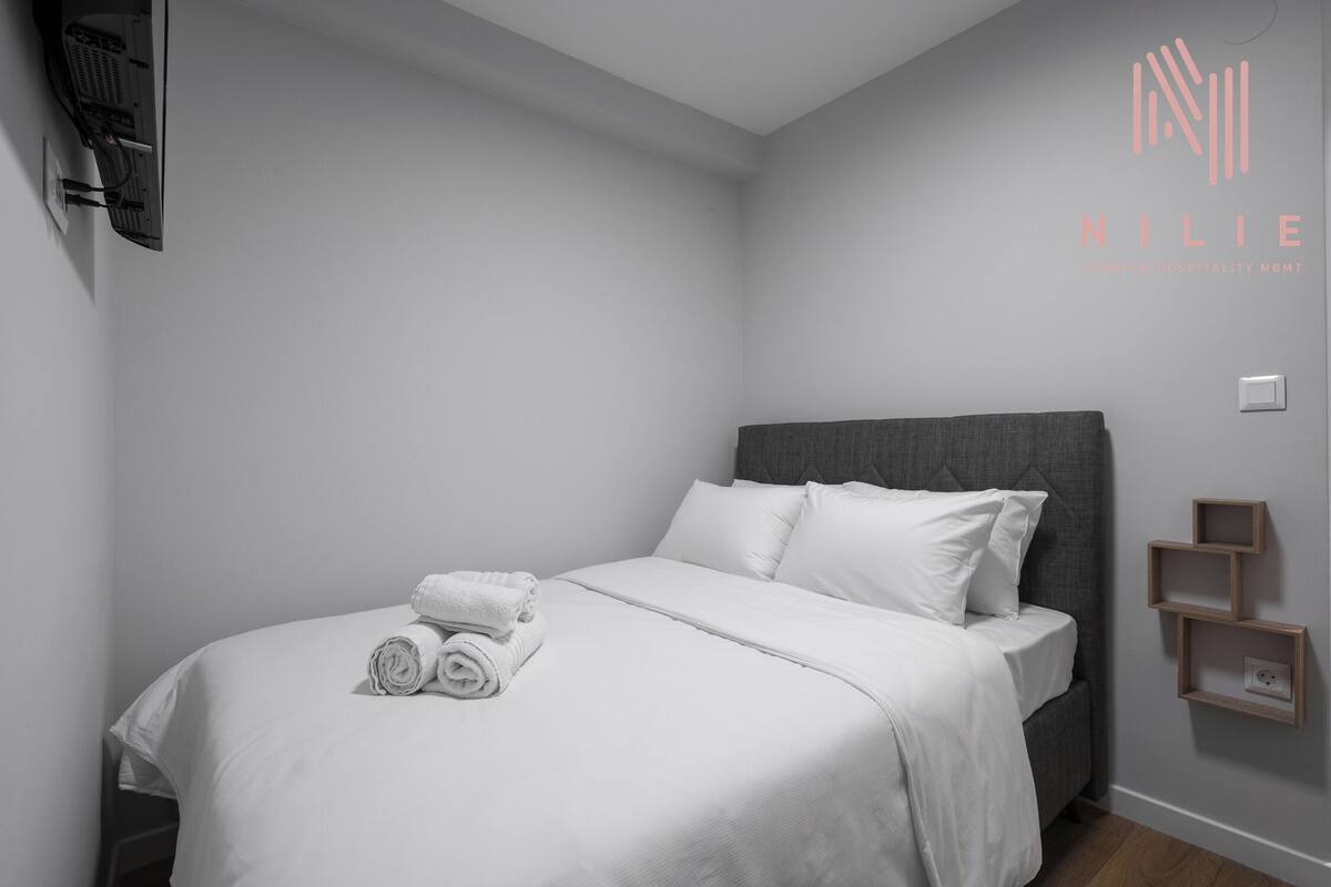 A neatly made bed with white linens and rolled towels, a grey headboard, and a wall-mounted shelf unit.