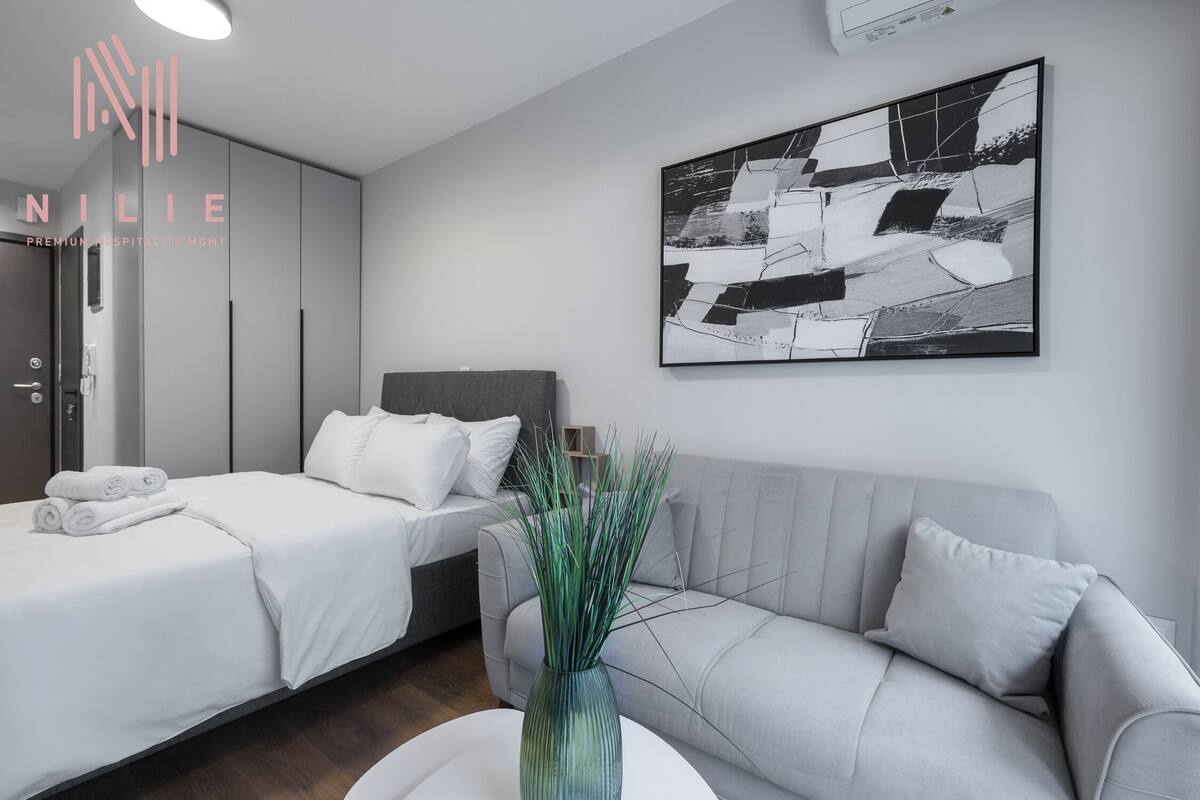 A modern hotel room with a bed, a sofa, a vase with green plants, and a black and white abstract painting on the wall.