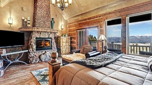 A spacious bedroom with a stone fireplace, a large bed, a television, and a balcony with a mountain view. - Spacious, Custom Built, Luxury Home Cabin with Hot Tub, Stunning Views,  (Angel Fire)