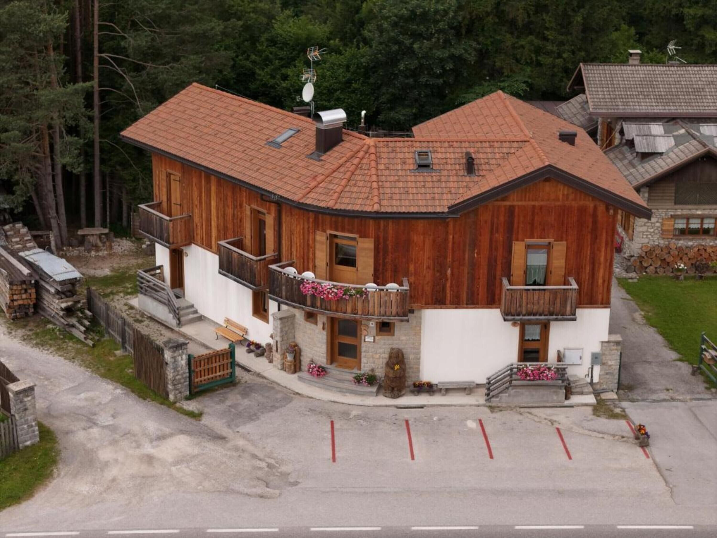 A two-story wooden house with a sloped roof, a balcony with flowers, and a stone wall entrance.