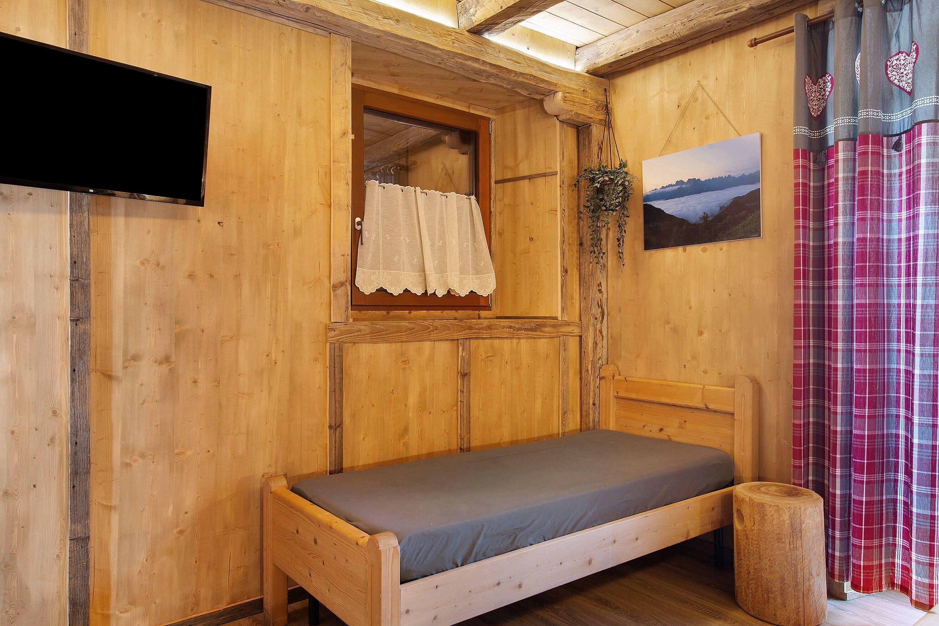 Una habitación de madera con cama, ventana con cortina, televisor montado y un cuadro en la pared.