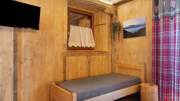 Una habitación de madera con cama, ventana con cortina, televisor montado y un cuadro en la pared.