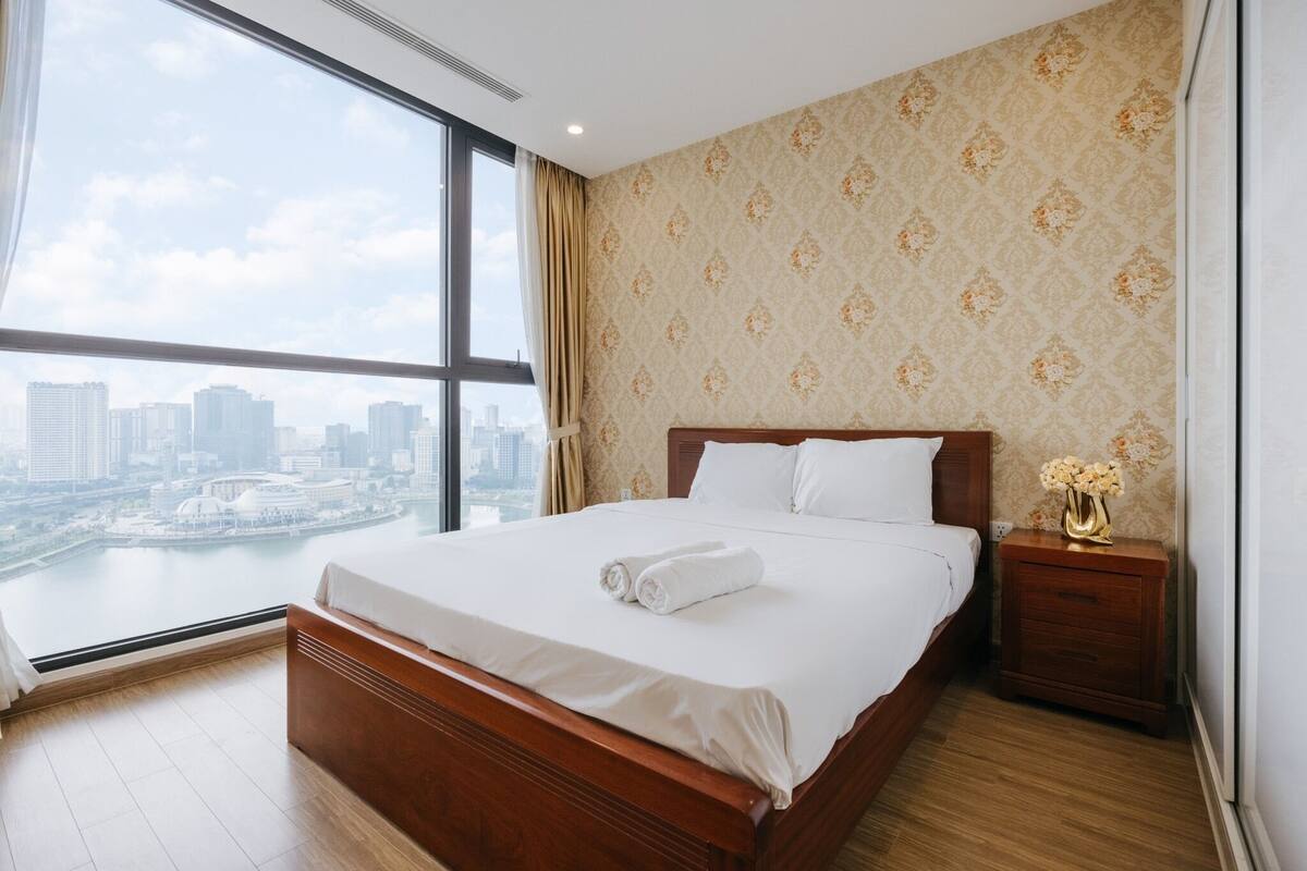 Ein Hotelzimmer mit einem großen Bett, einem hölzernen Kopfteil, einem Nachttisch mit Blumenvase und Blick auf eine Stadtkulisse durch ein großes Fenster.