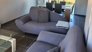 A modern living room with a grey sofa, a glass coffee table, and a dining area with a table and chairs.