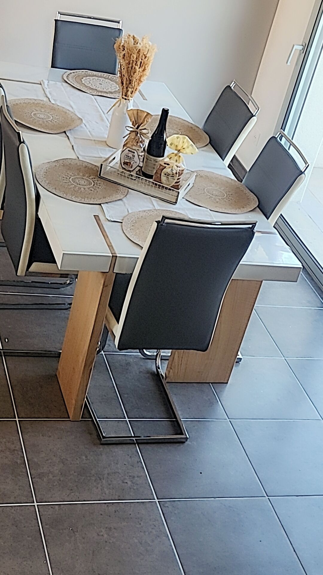 A dining area with a white table set for six, featuring a bottle of wine, a vase with dried flowers, and a menu. Chairs are upholstered in black and wood.
