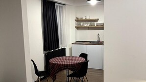 A compact living space with a dining area featuring a table and chairs, a kitchenette with a sink and shelves, and a window with curtains. - Gudauri - Apartment - Sleeps 4 - Pool - Parking (Kazbegi)