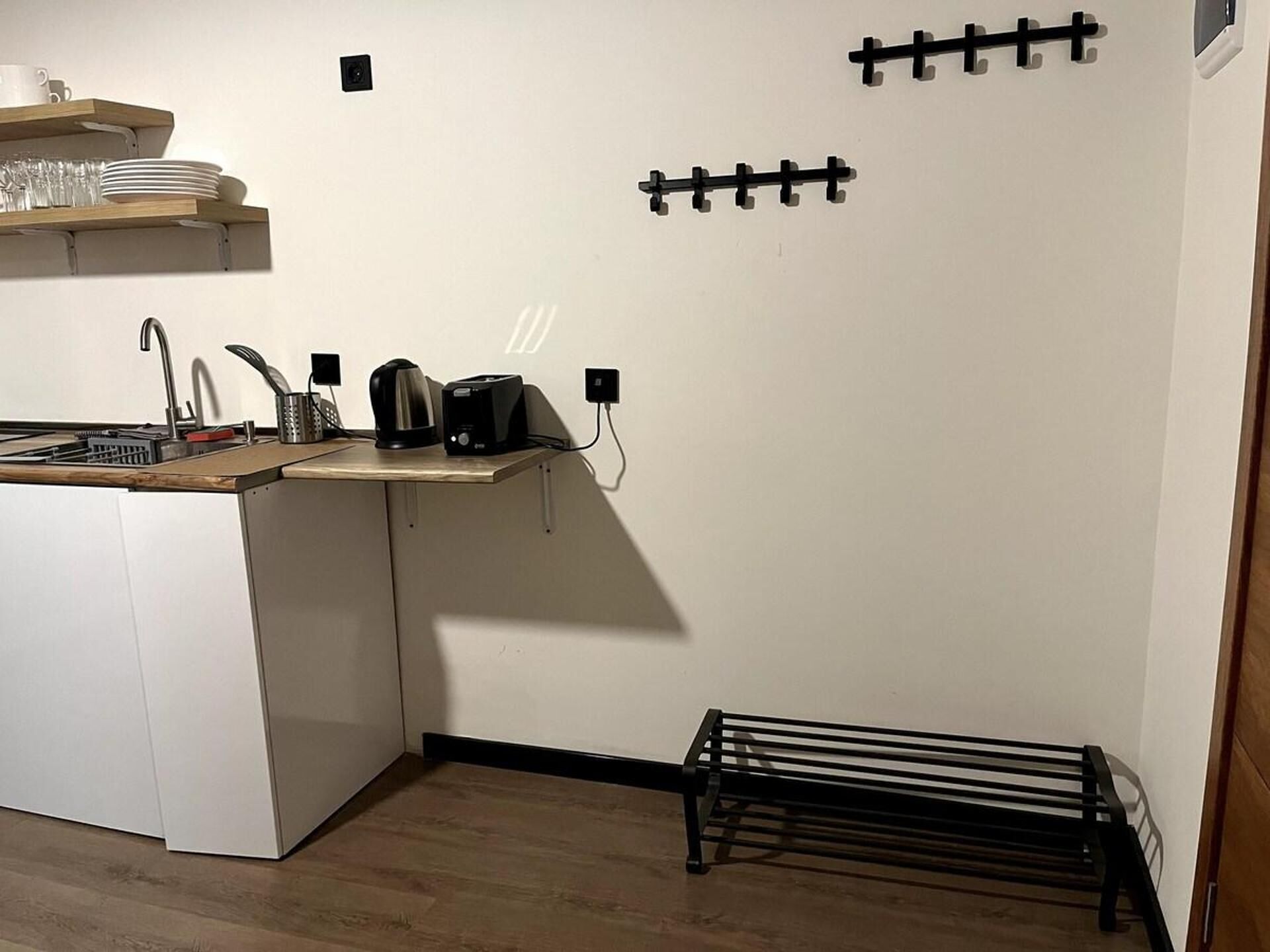 A compact kitchen with a sink, countertop, and a coffee maker. There is a wooden shelf above the sink holding plates and glasses.