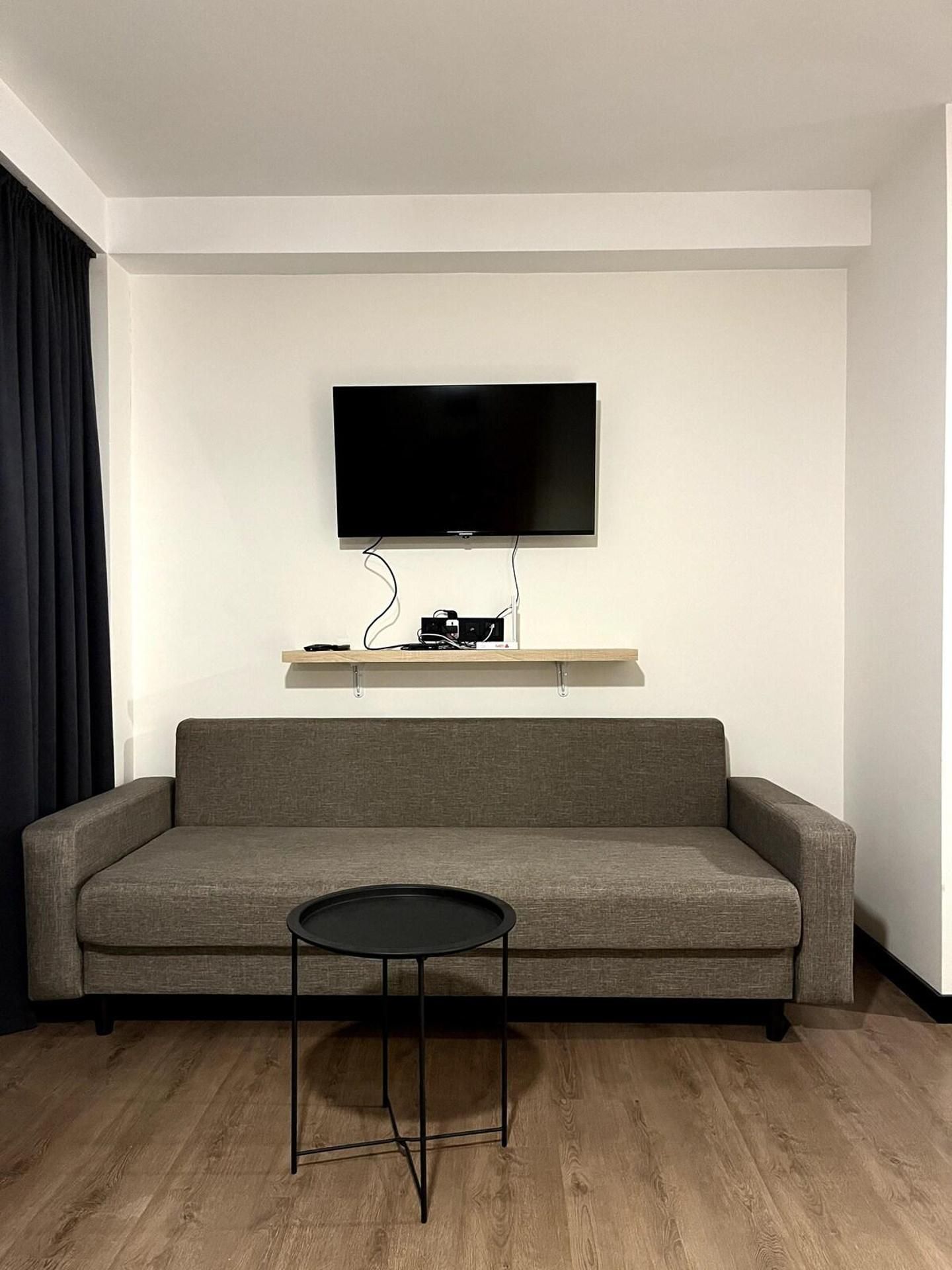 Un saló modern amb un sofà gris, una taula petita negra i un televisor pla fixat sobre una estant de fusta.