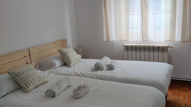 A hotel room with two beds, a wooden headboard, white bedding, a radiator, and sheer curtains. - 1A135 Comodo Apto 3 dormitorios (Gijón)