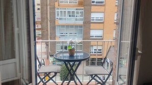 A balcony with a round table and two chairs, a potted plant, and a view of a multi-story building. - 1A135 Comodo Apto 3 dormitorios (Gijón)