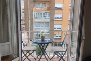 Ein Balkon mit rundem Tisch, zwei Stühlen, einem Blumentopf und Blick auf ein mehrgeschossiges Gebäude.
