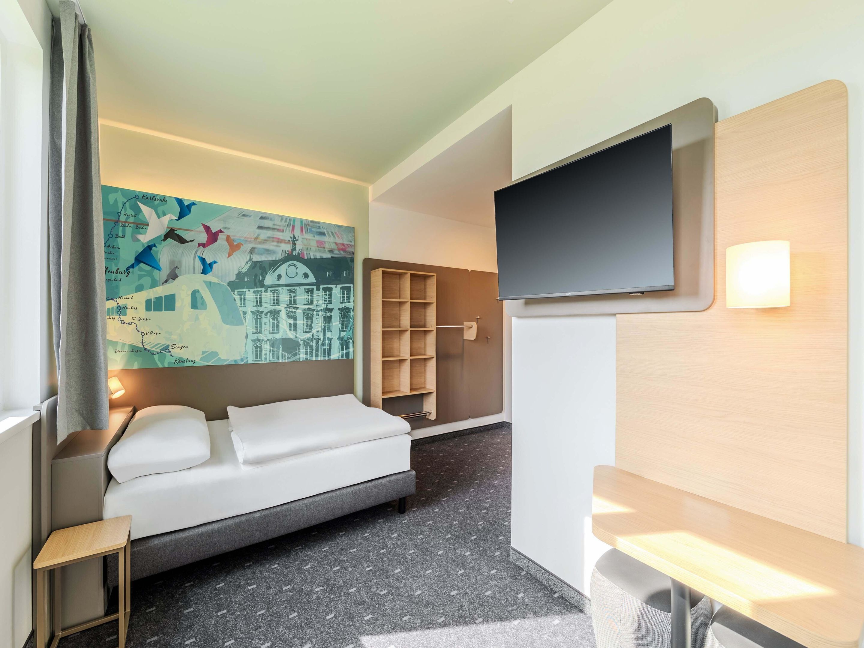 A modern hotel room with a bed, a flat-screen TV, a wooden side table, a small wooden stool, and a wall-mounted shelf.