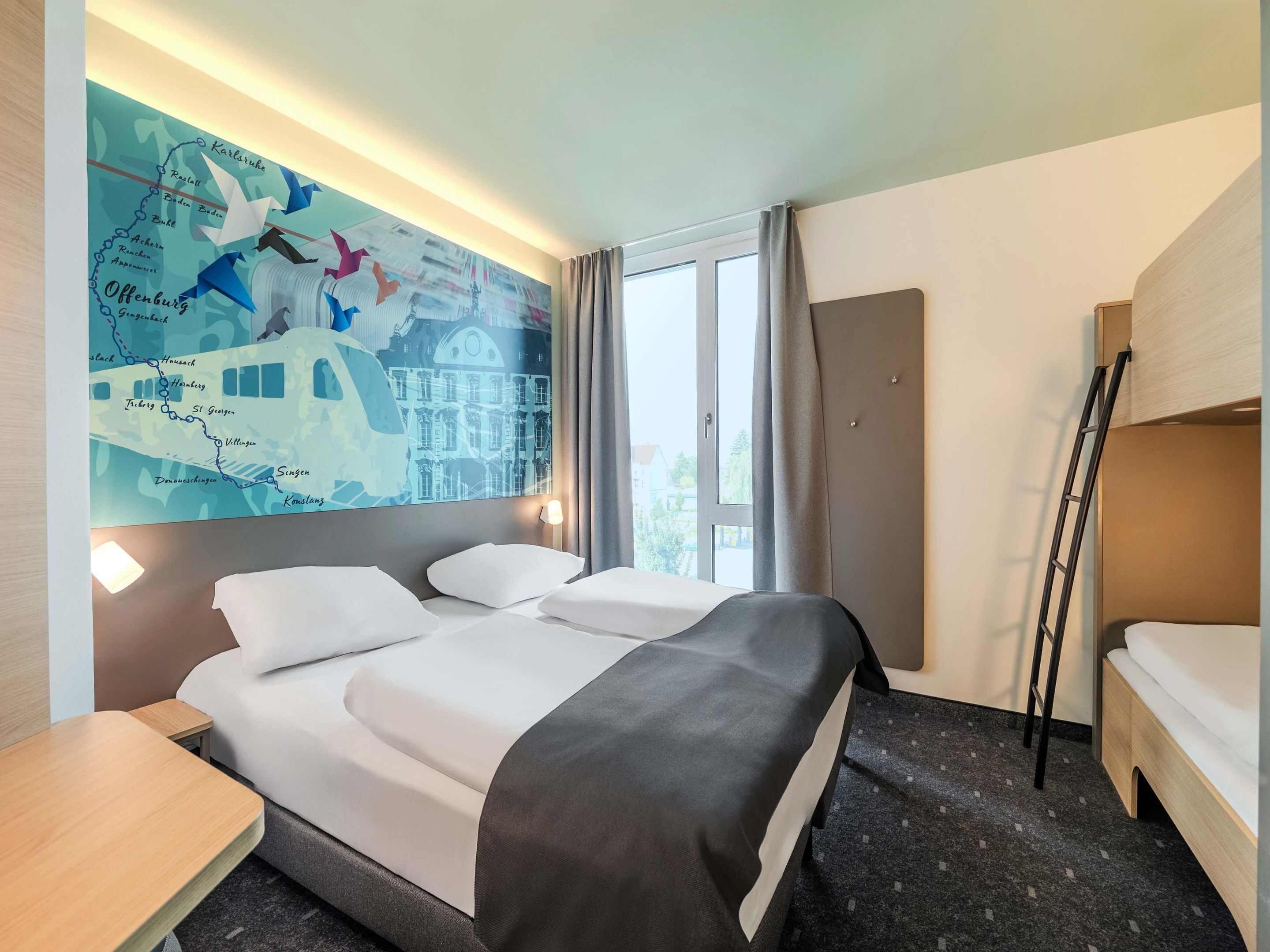 Quarto de hotel com uma cama grande, uma escrivaninha, um mapa na parede e uma janela com cortinas.