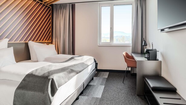 Ein modernes Hotelzimmer mit Bett, Schreibtisch, Stuhl und einem großen Fenster mit Blick auf die Stadt.
