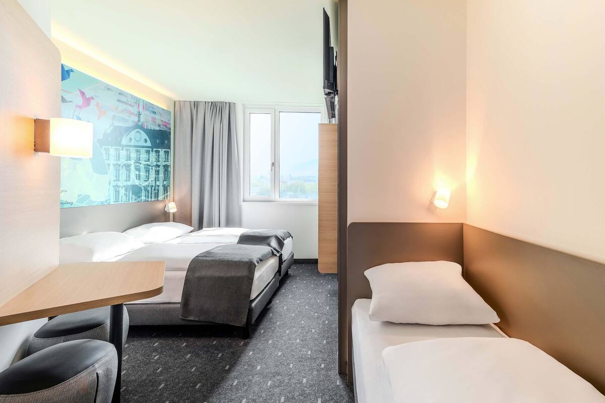 A hotel room with two beds, a desk, and a chair. There is a painting on the wall and a window with curtains.