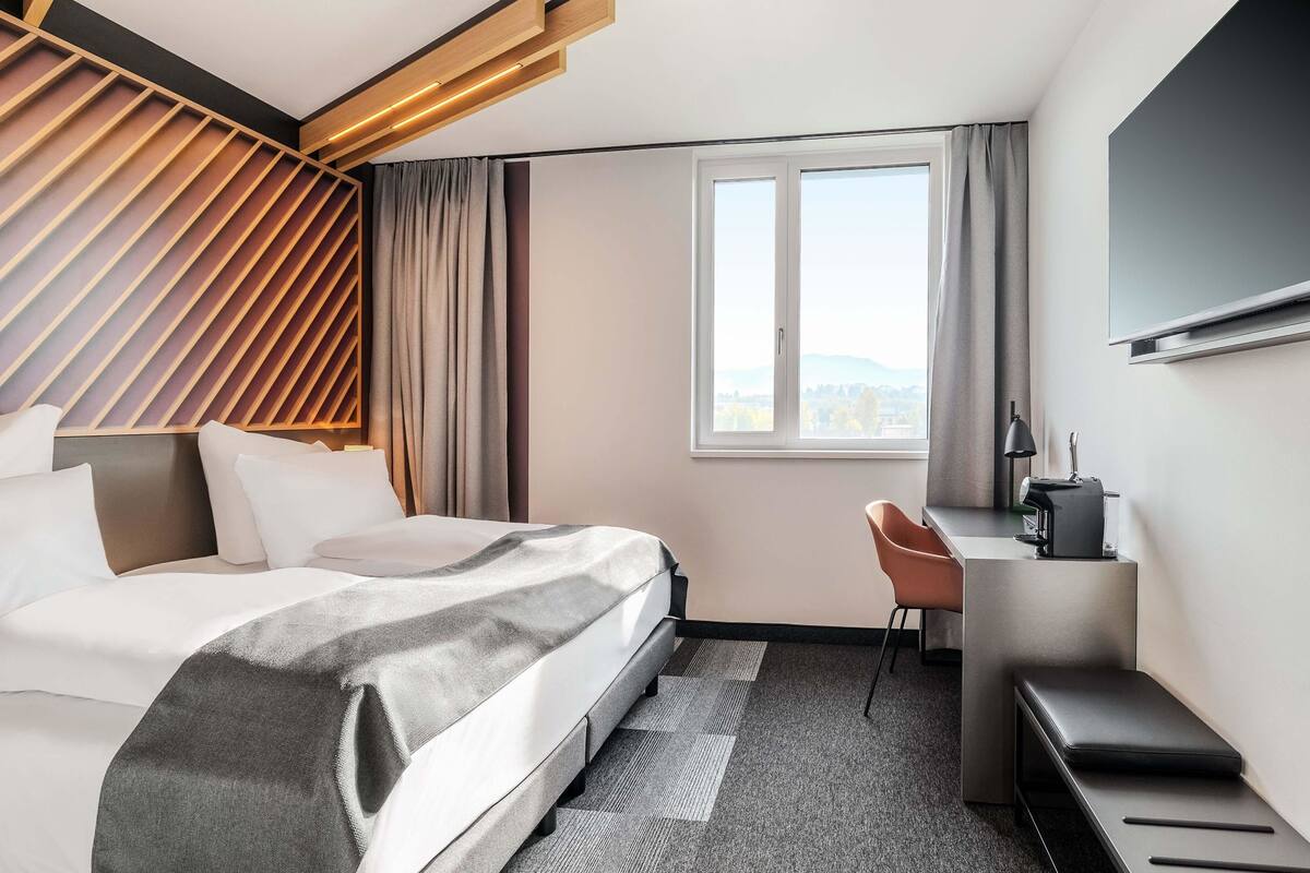 A modern hotel room with a bed, a desk, a chair, and a large window offering a city view.