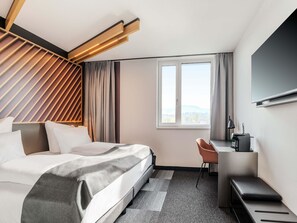 A modern hotel room with a bed, a desk, a chair, and a large window offering a city view.