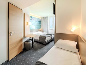 Habitación de hotel con cama, una mesita y una silla. Tiene una ventana con cortinas, un televisor montado en la pared y una lámpara de pared.