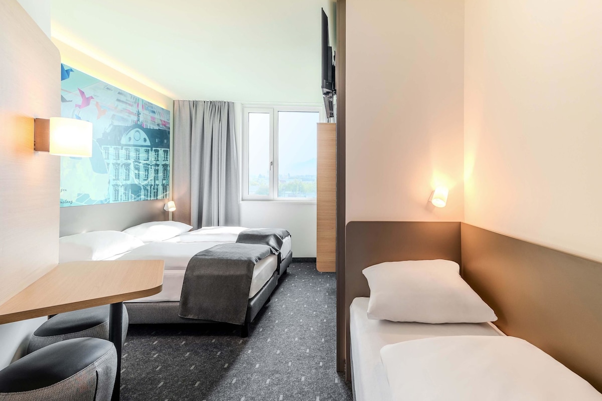 Een hotelkamer met twee bedden, een bureau en een stoel. Er hangt een schilderij aan de muur en er is een raam met gordijnen.