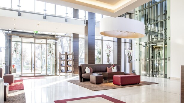 A spacious lobby with a modern design, featuring a large seating area with a sofa and ottoman, a reception desk, and a large window offering a view of the outside.