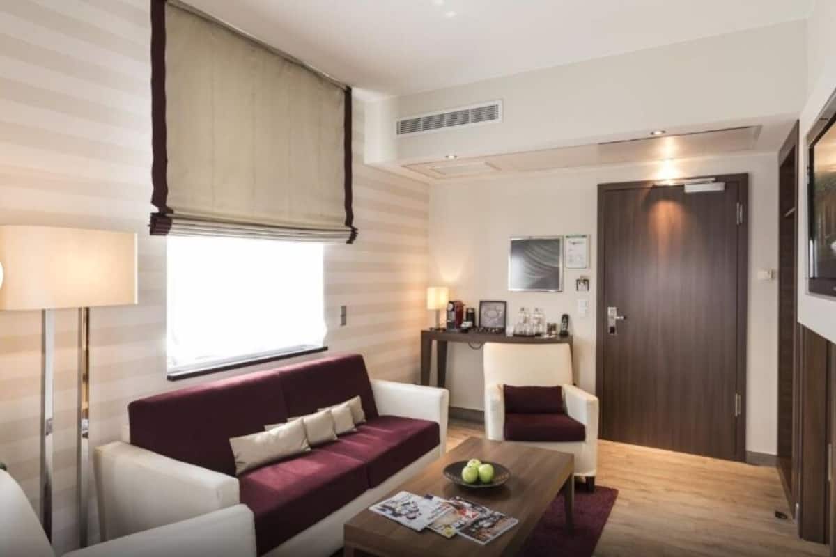 A modern hotel room with a sofa, armchairs, a coffee table, and a large window with a blind.