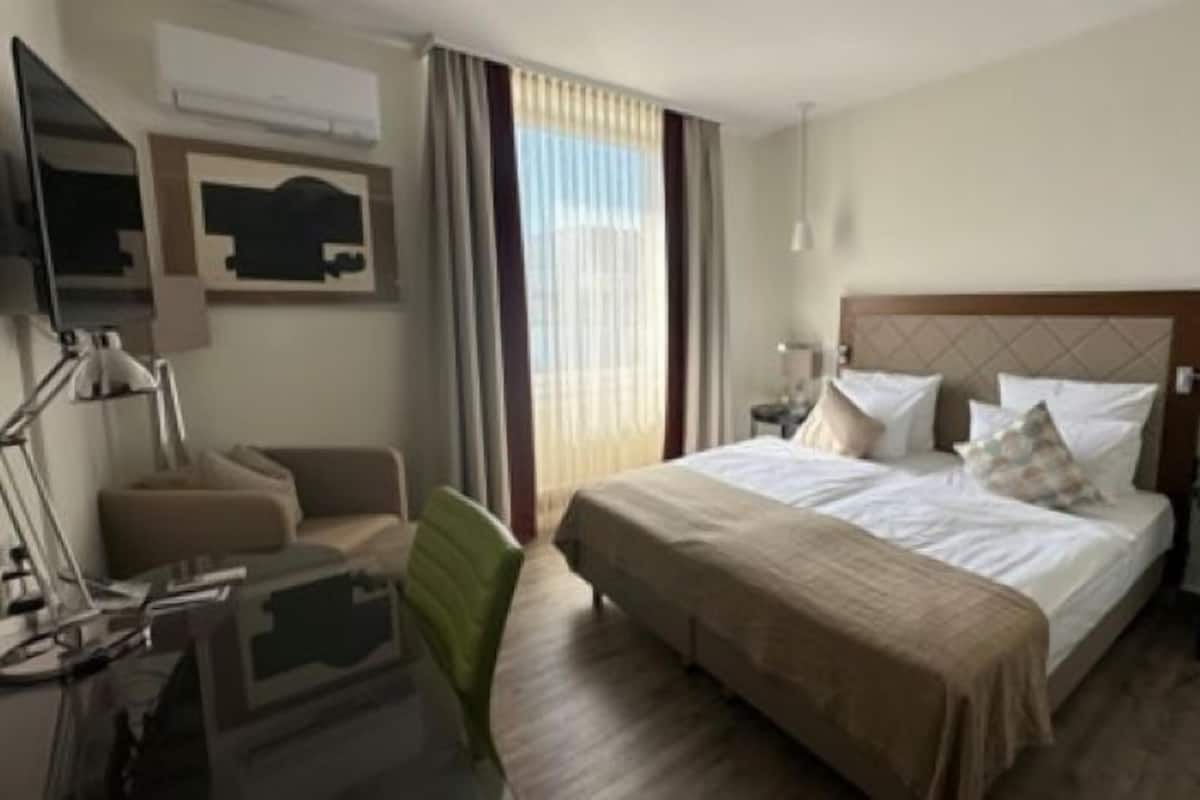 A hotel room with a large bed, two bedside tables, a desk, a chair, a lamp, and a window with curtains.